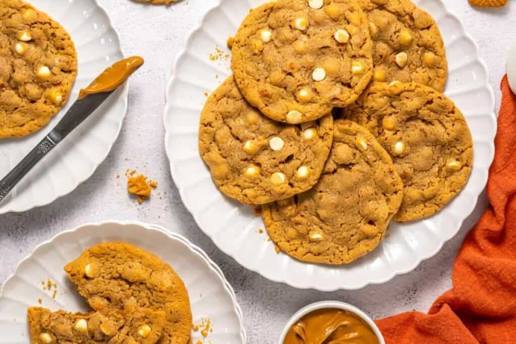 A plate of peanut butter cookies with white chocolate chips is shown on a white surface, alongside a knife, a small bowl of peanut butter, and an orange napkin.