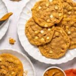 A plate of cookies with white chocolate chips is shown on a white surface, alongside a knife, a small bowl of Biscoff cookie butter, and an orange napkin.