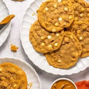 A plate of cookies with white chocolate chips is shown on a white surface, alongside a knife, a small bowl of peanut butter, and an orange napkin.