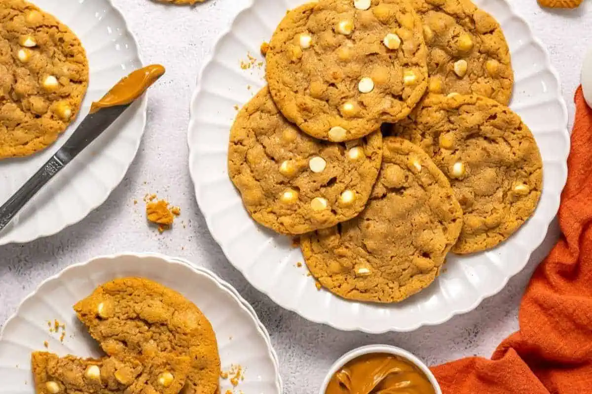 A plate of large peanut Copycat Crumbl Biscoff cookies with white chocolate chips, surrounded by a knife, a small bowl of peanut butter, and an orange napkin.