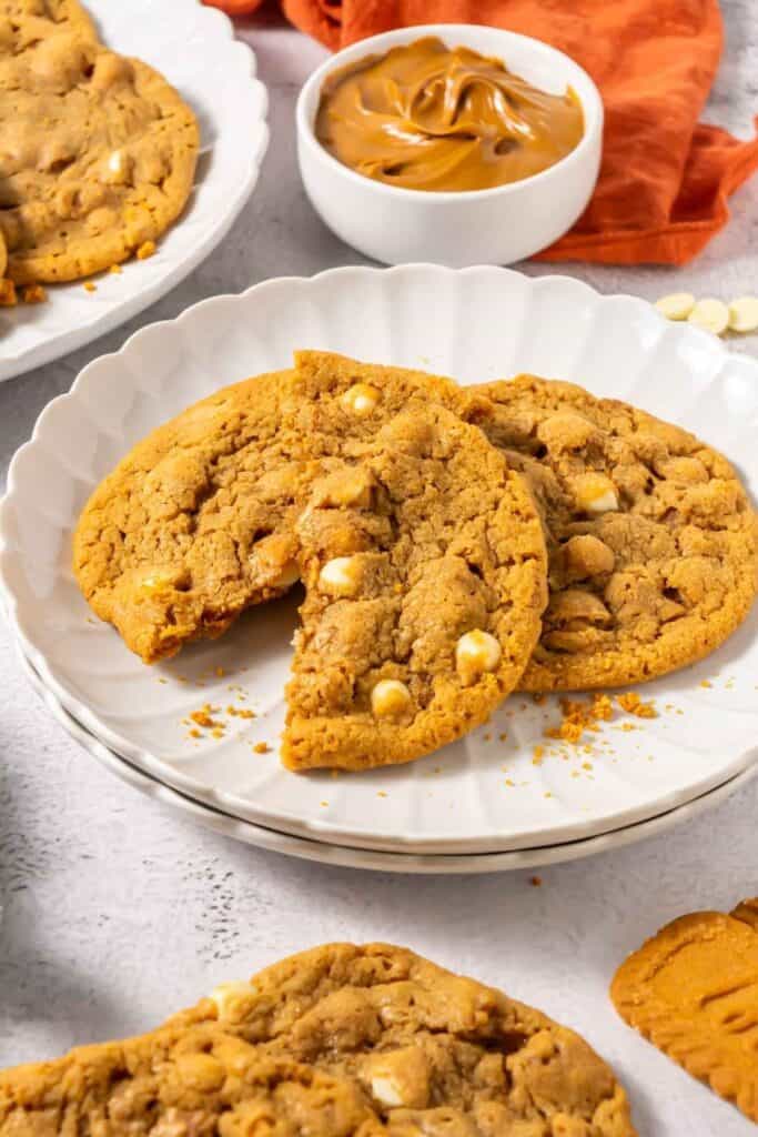 Two peanut butter cookies with white chocolate chips on a white plate, one with a bite taken out. A bowl of peanut butter and more cookies are in the background.
