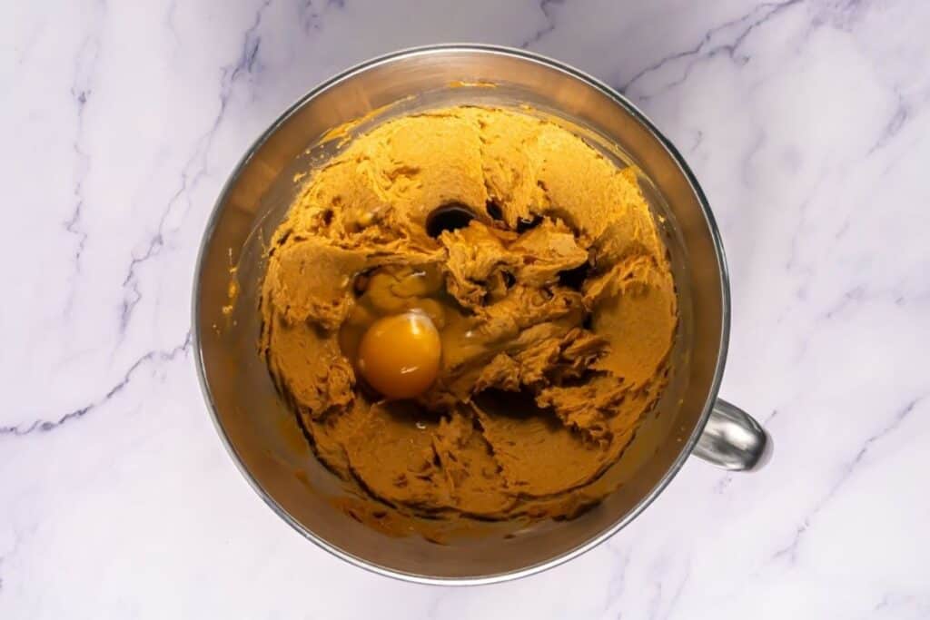 A metal mixing bowl containing brown cookie dough with a cracked raw egg on top, placed on a white marble surface.