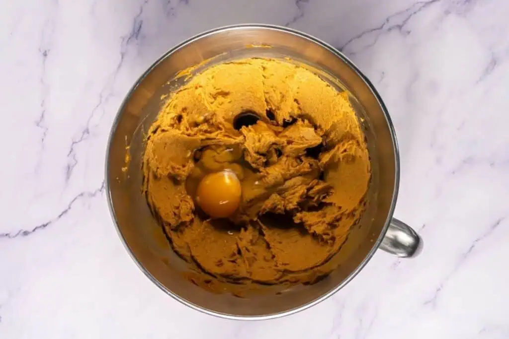 A metal mixing bowl containing brown cookie dough with a cracked raw egg on top, placed on a white marble surface.