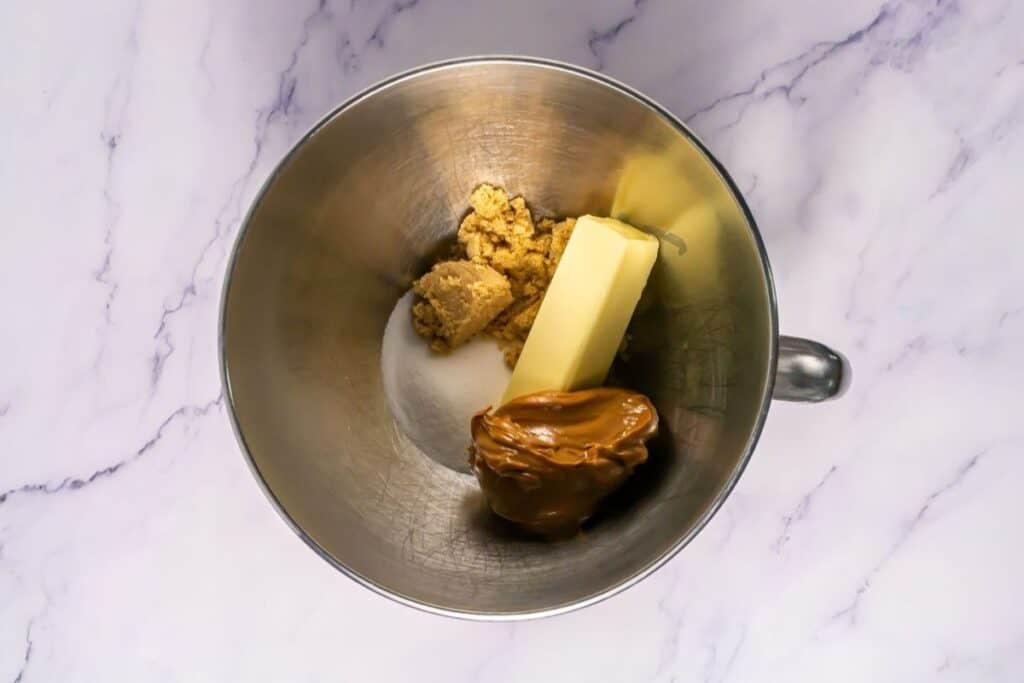 A mixing bowl containing brown sugar, white sugar, a stick of butter, and a large dollop of peanut butter on a marble surface.