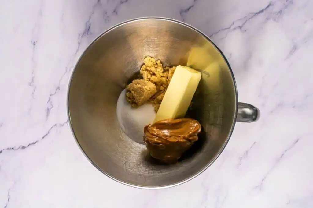 A mixing bowl containing brown sugar, white sugar, a stick of butter, and a large dollop of peanut butter on a marble surface.