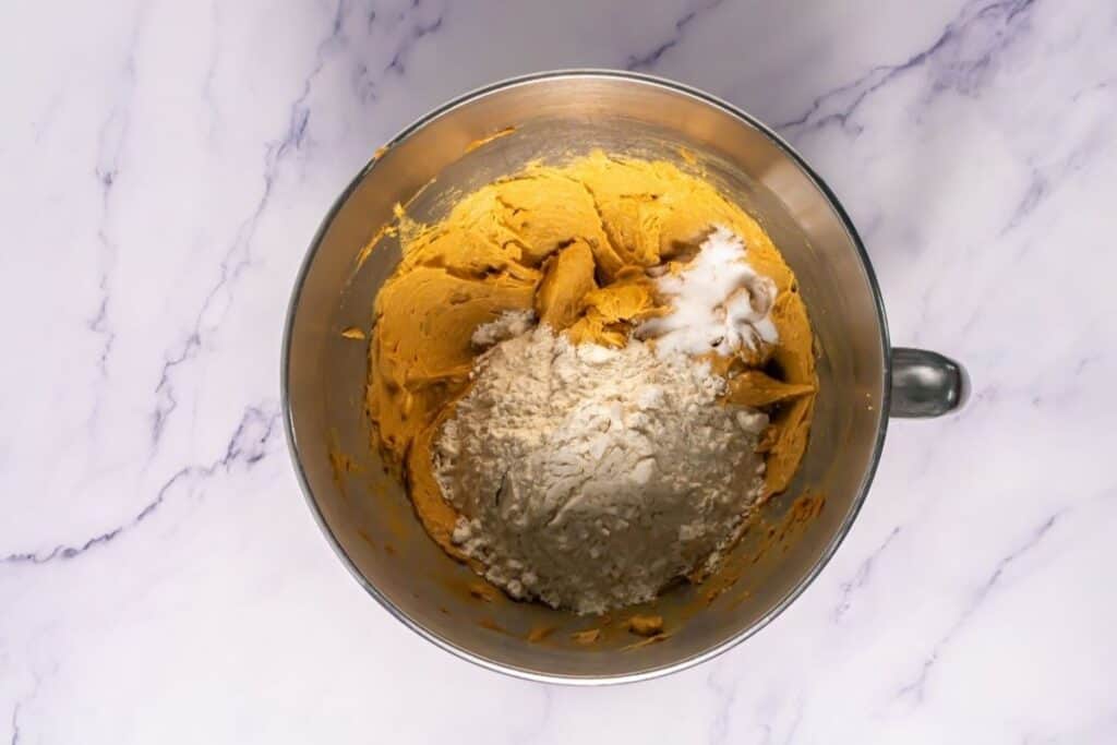 A metal mixing bowl containing partially mixed cookie dough with flour, brown dough, and white granules visible, on a white marble surface.