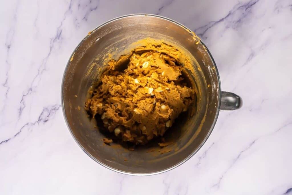 A metal mixing bowl containing cookie dough with white chocolate chips on a white marble surface.