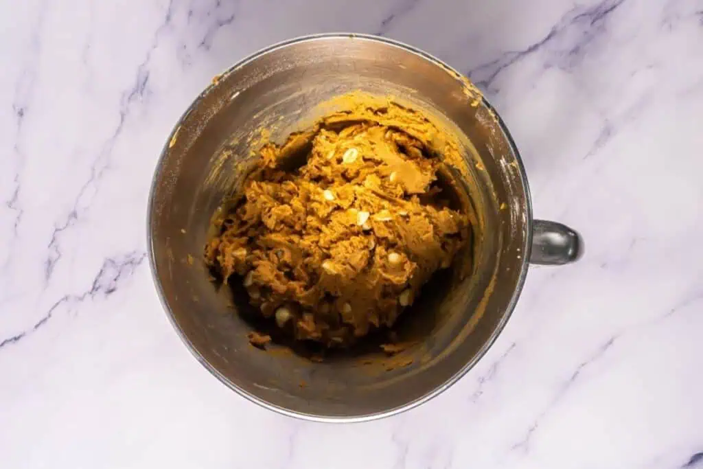 A metal mixing bowl containing cookie dough with white chocolate chips on a white marble surface.