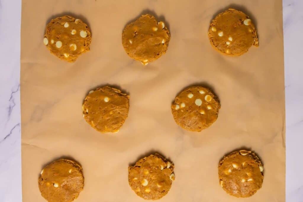 Nine unbaked cookies with white chocolate chips are arranged in three rows on a parchment-lined baking sheet.