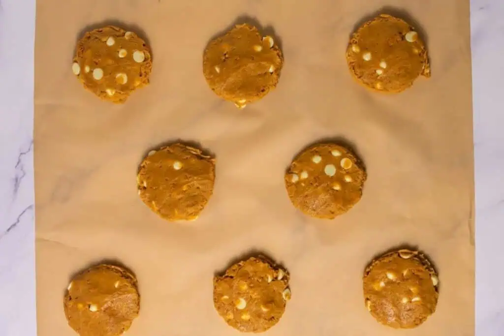 Nine unbaked cookies with white chocolate chips are arranged in three rows on a parchment-lined baking sheet.