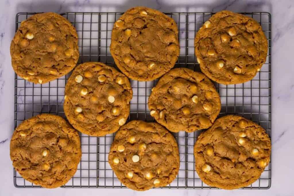 Eight large cookies with white chocolate chips cooling on a metal wire rack, placed on a white marble surface.