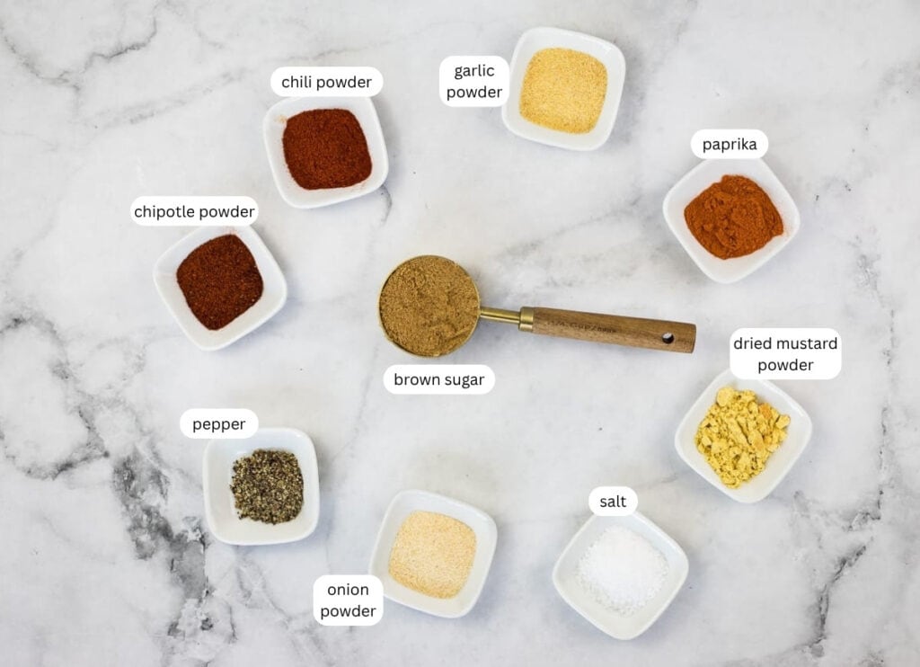 Nine small bowls with labeled spices and seasonings, including brown sugar, pepper, salt, paprika, chili powder, chipotle powder, onion powder, garlic powder, and dried mustard powder.