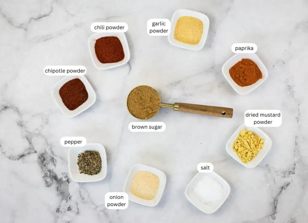 Nine small bowls with labeled spices and seasonings, including brown sugar, pepper, salt, paprika, chili powder, chipotle powder, onion powder, garlic powder, and dried mustard powder.