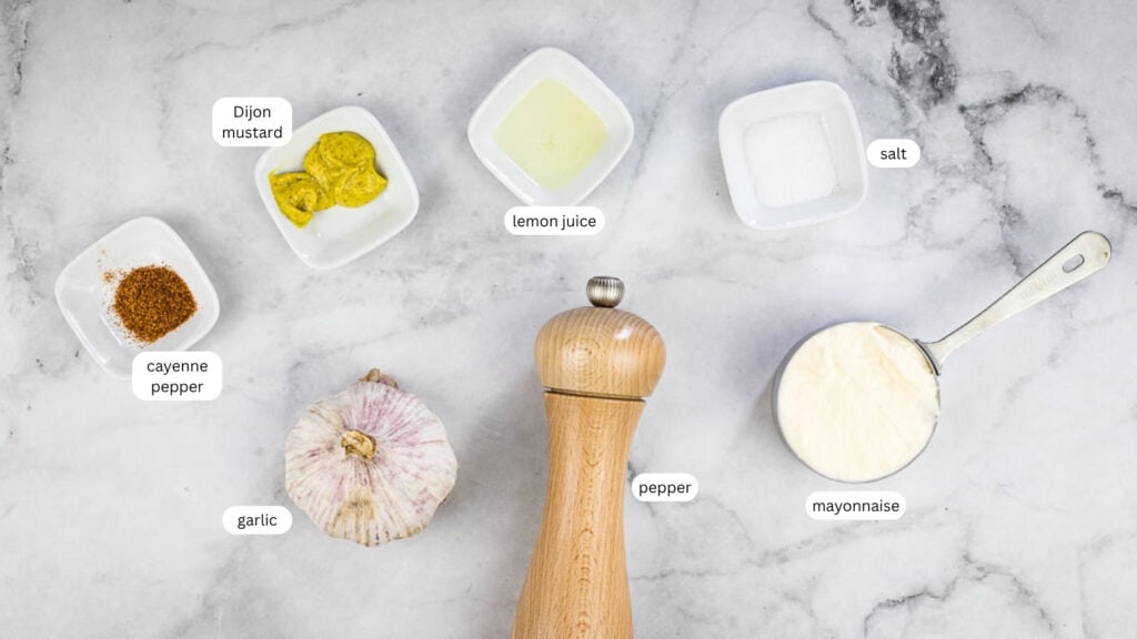 Ingredients for a sauce arranged on a marble surface: cayenne pepper, garlic, Dijon mustard, lemon juice, salt, pepper grinder, and a measuring cup of mayonnaise.