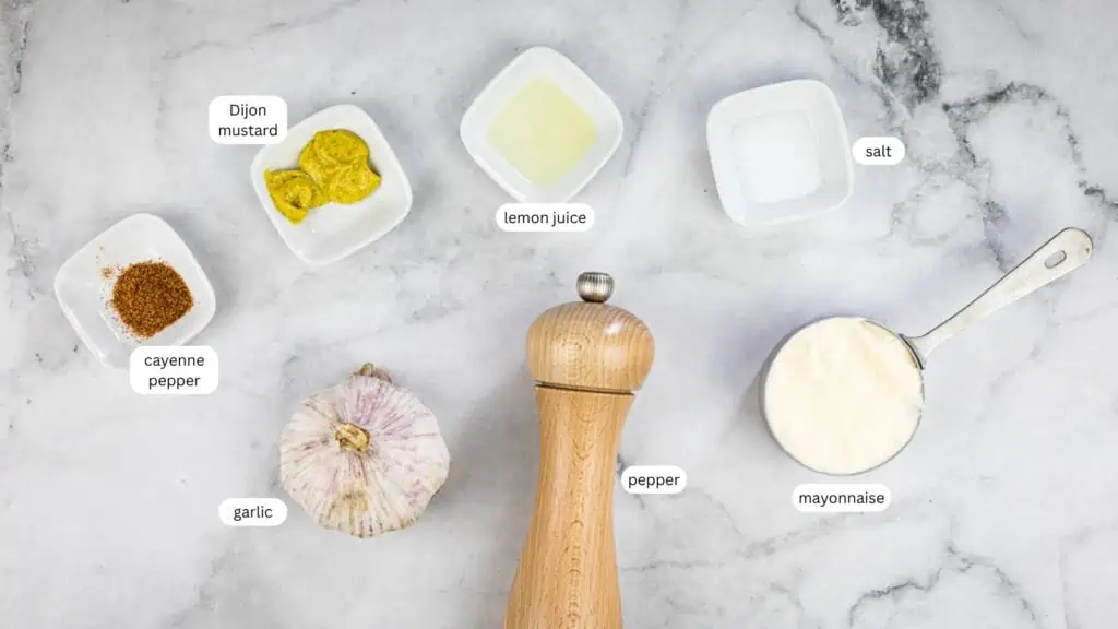 Ingredients for a sauce arranged on a marble surface: cayenne pepper, garlic, Dijon mustard, lemon juice, salt, pepper grinder, and a measuring cup of mayonnaise.