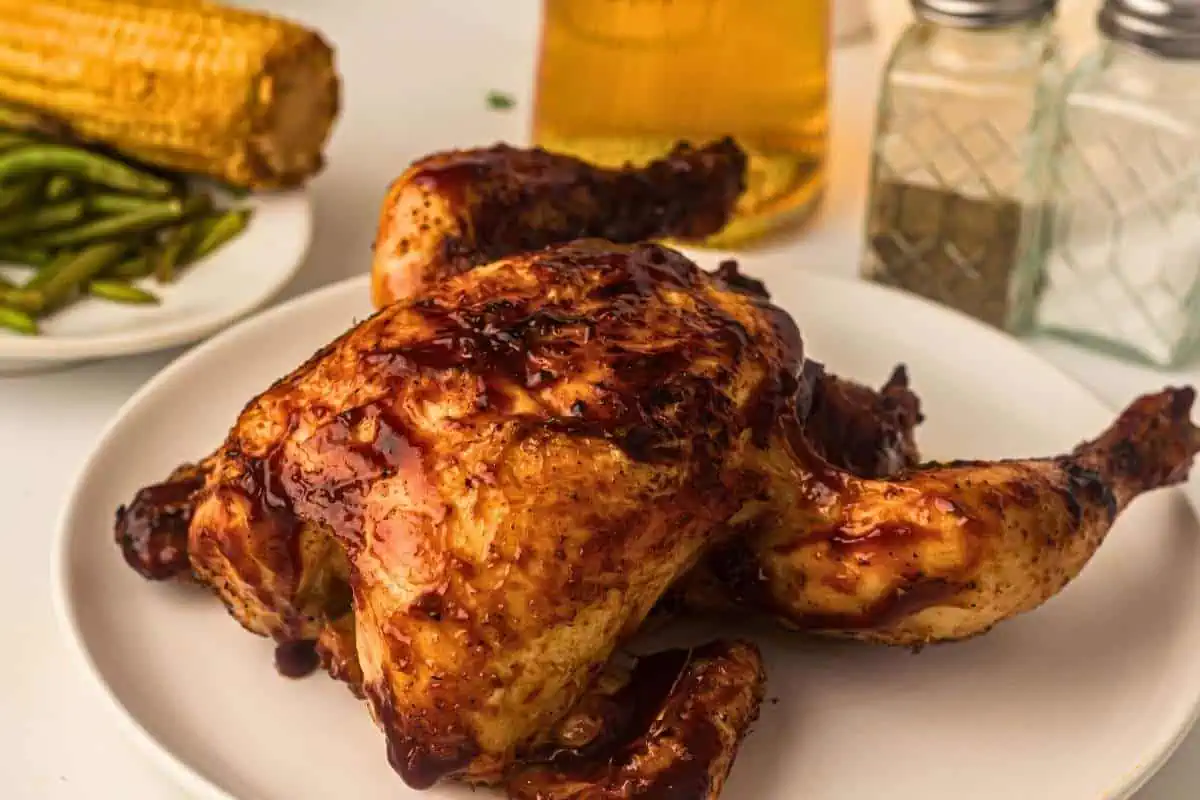 A roasted chicken with barbecue sauce on a white plate, with green beans, corn on the cob, a glass of beer, and salt and pepper shakers in the background.