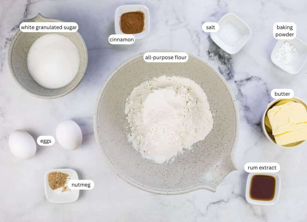 A flat lay of baking ingredients: all-purpose flour, eggs, butter, white granulated sugar, cinnamon, nutmeg, rum extract, baking powder, and salt in separate bowls on a white surface.