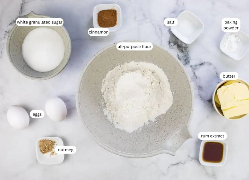 A flat lay of baking ingredients: all-purpose flour, eggs, butter, white granulated sugar, cinnamon, nutmeg, rum extract, baking powder, and salt in separate bowls on a white surface.