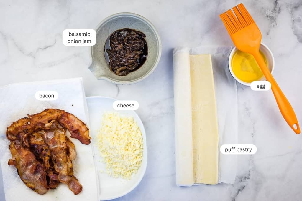 Ingredients for a recipe are laid out: cooked bacon, shredded cheese, balsamic onion jam in a bowl, two puff pastry sheets, a bowl of beaten egg, and an orange pastry brush.