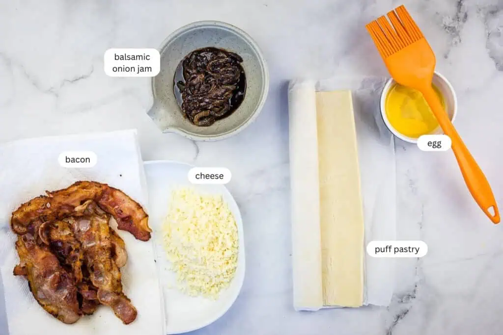 Ingredients for a recipe are laid out: cooked bacon, shredded cheese, balsamic onion jam in a bowl, two puff pastry sheets, a bowl of beaten egg, and an orange pastry brush.
