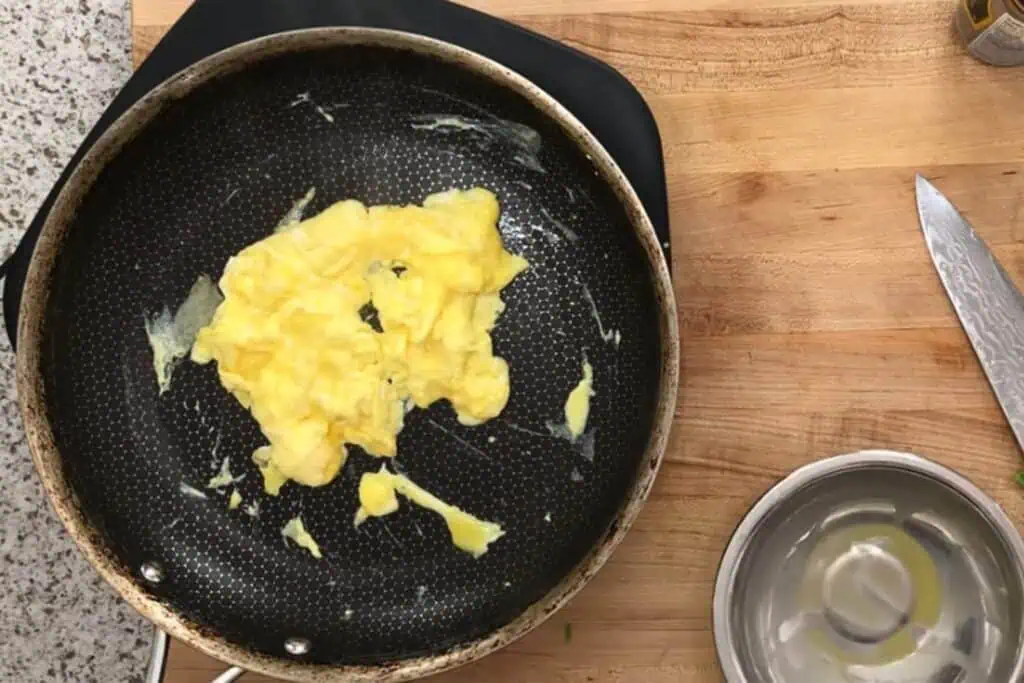 A small portion of scrambled eggs cooking in a nonstick frying pan on a wooden countertop, with a knife and metal bowl nearby.