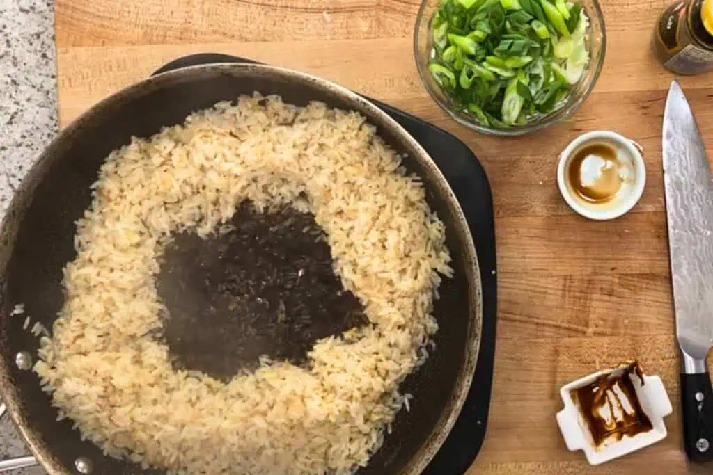 Fried rice in a pan with a sauce in the center, surrounded by bowls of chopped green onions, sauces, and a knife on a wooden countertop.
