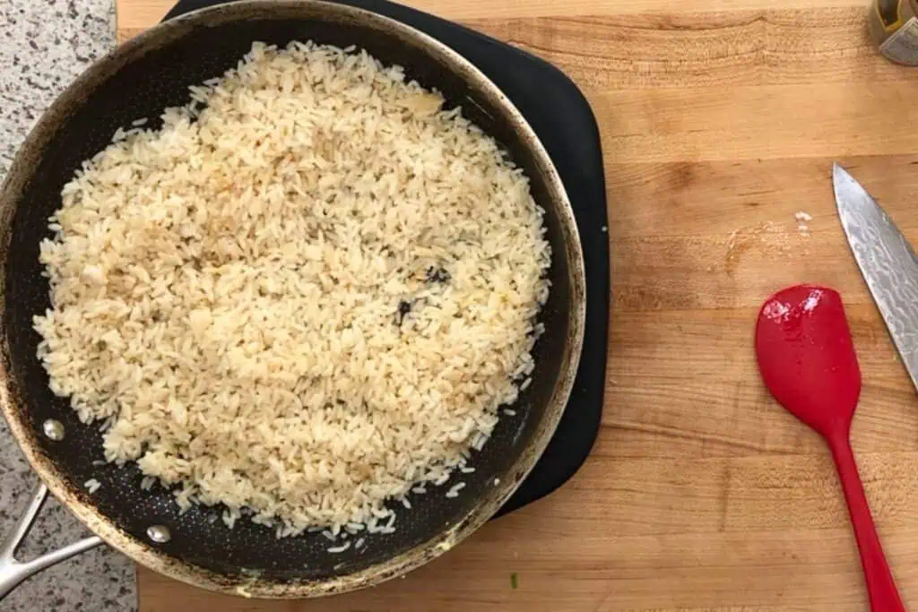 A frying pan filled with cooked rice sits on a wooden countertop beside a red spatula and a knife.
