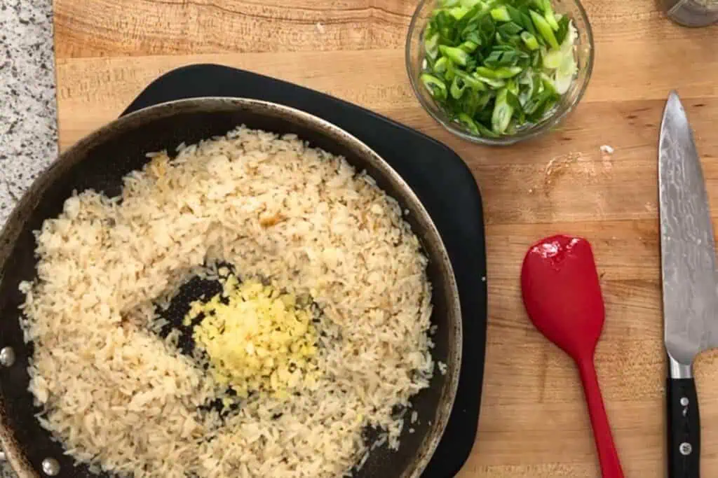 A frying pan with cooked rice and minced garlic sits on a wooden countertop, next to a bowl of chopped green onions, a chef’s knife, and a red spatula.