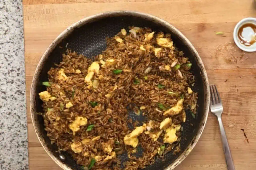 A frying pan with fried rice, scrambled eggs, and scallions sits on a wooden cutting board next to a fork and a small dish with sauce residue.