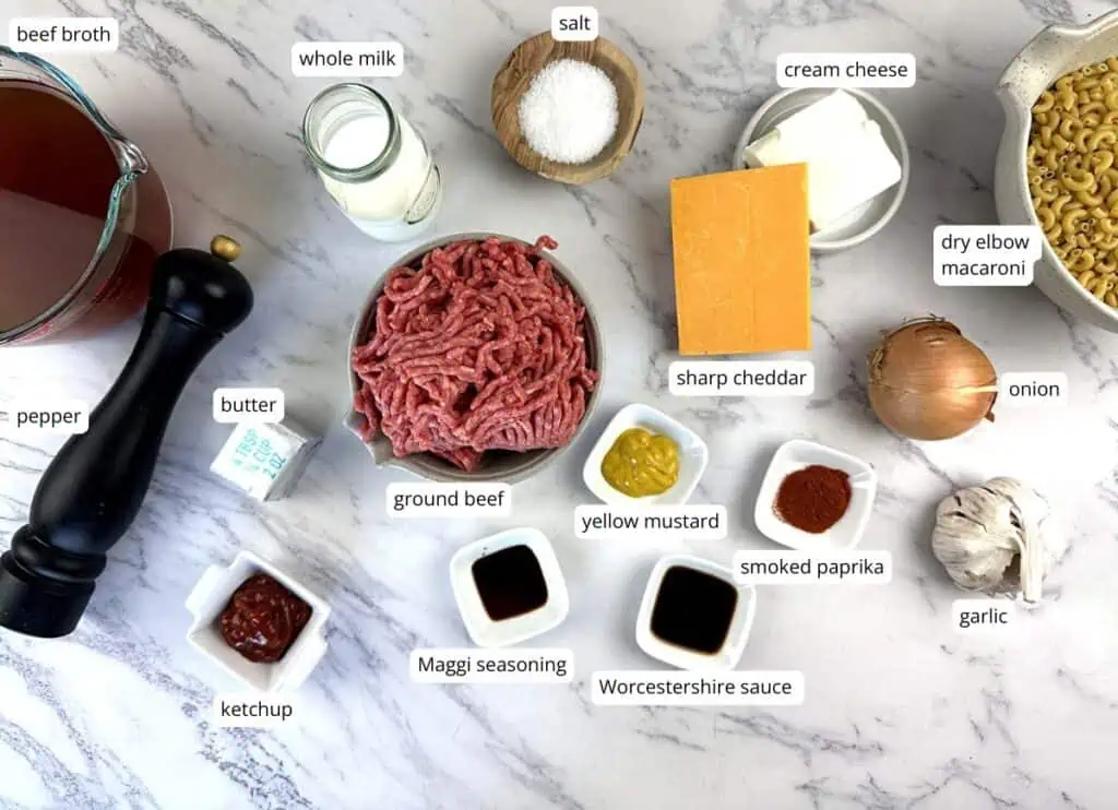 Ingredients for a beef macaroni dish arranged on a marble surface, including ground beef, pasta, cheese, milk, onion, garlic, spices, ketchup, and seasonings.
