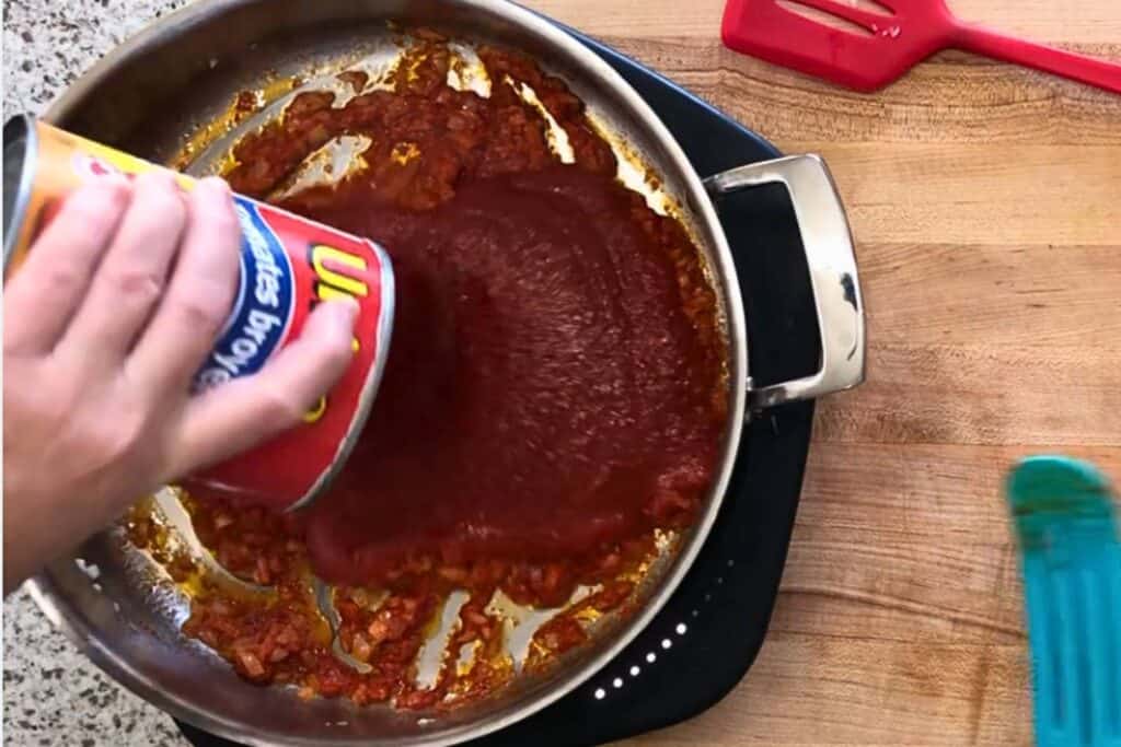 A person pours canned tomato sauce into a pan with sautéed ingredients on a stovetop; a red spatula and blue utensil are nearby on a wooden countertop.