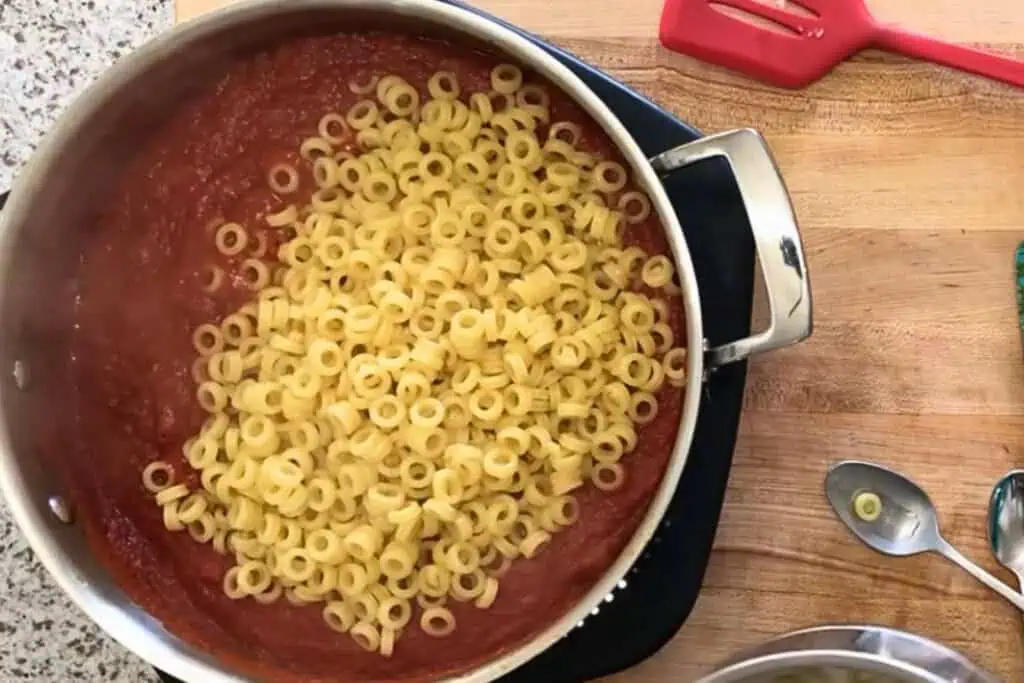 A pan with tomato sauce and uncooked anelli pasta on a wooden countertop, beside a red spatula and a spoon with a piece of pasta on it.