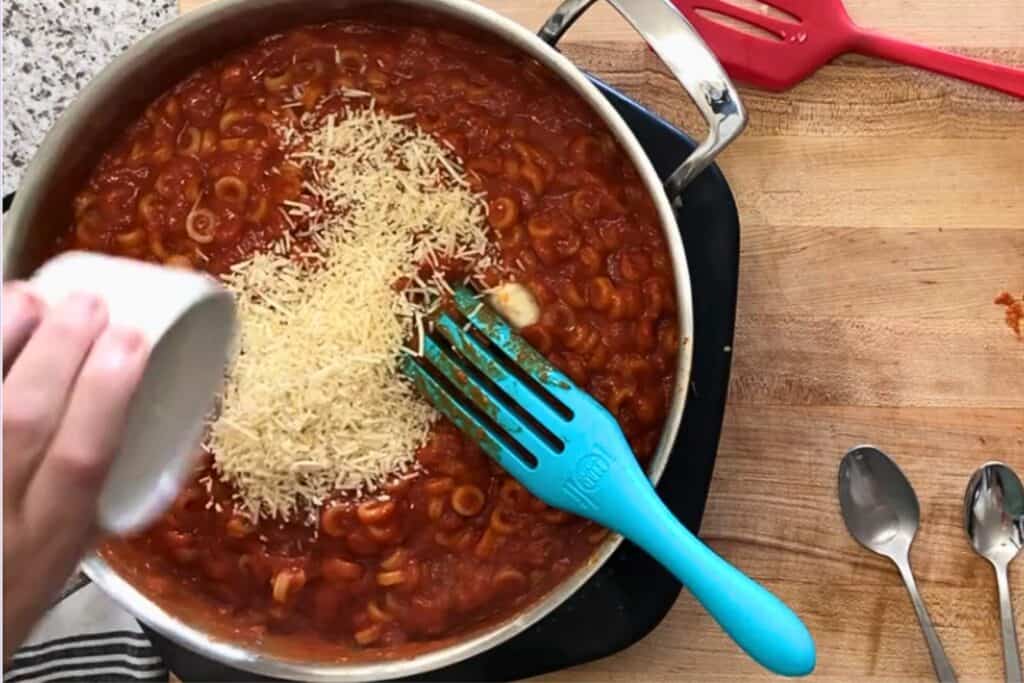 A hand sprinkles shredded cheese into a pan of tomato pasta sauce with o-shaped pasta on a stovetop, with utensils and spoons nearby on a wooden counter.