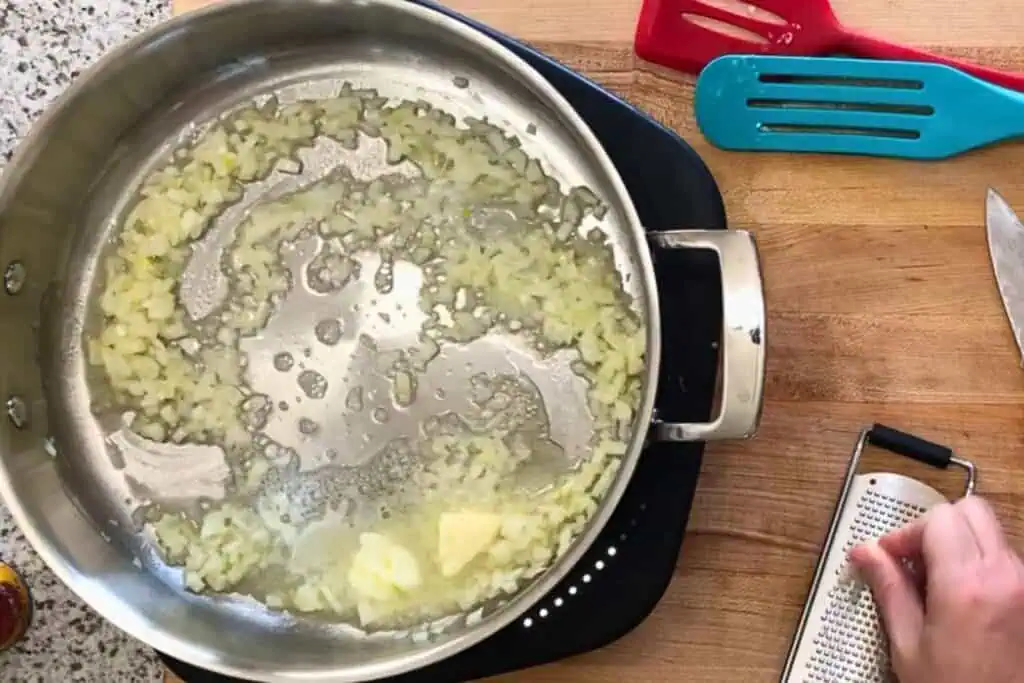 Chopped onions are sautéing in a stainless steel pan with butter on a stovetop; a hand is grating cheese on a wooden surface nearby.