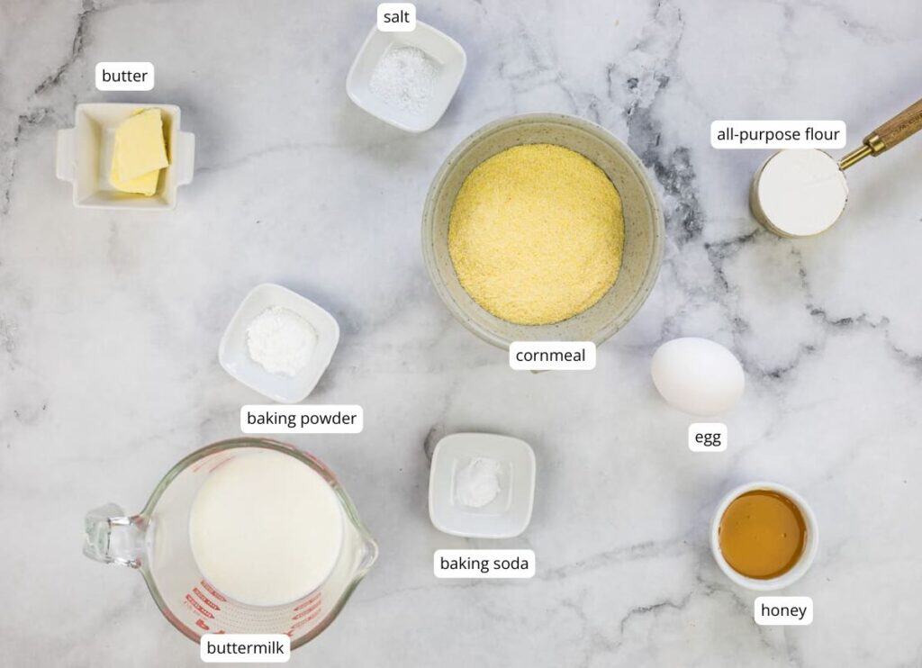 Ingredients for cornbread are arranged on a marble surface, including butter, salt, cornmeal, flour, an egg, baking powder, baking soda, honey, and buttermilk.