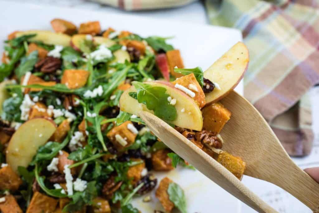 A close-up of a wooden fork holding a bite of salad with apple slices, arugula, pecans, feta cheese, and roasted sweet potatoes. The salad is on a white plate in the background.