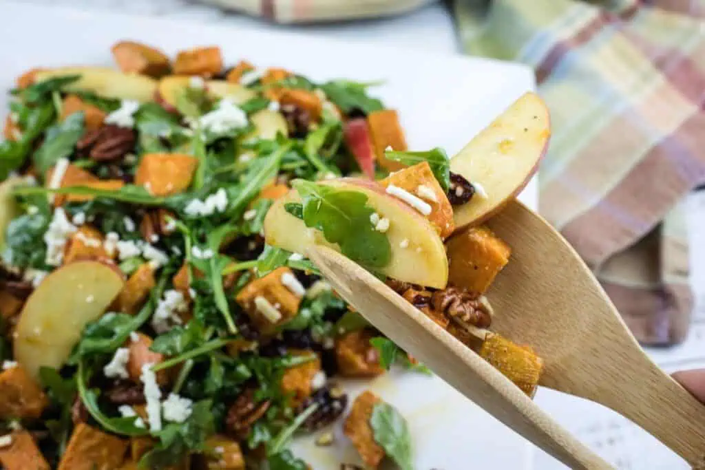 A close-up of a wooden fork holding a bite of salad with apple slices, arugula, pecans, feta cheese, and roasted sweet potatoes. The salad is on a white plate in the background.