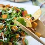A close-up of a wooden fork holding a bite of salad with apple slices, arugula, pecans, feta cheese, and roasted sweet potatoes. The salad is on a white plate in the background.