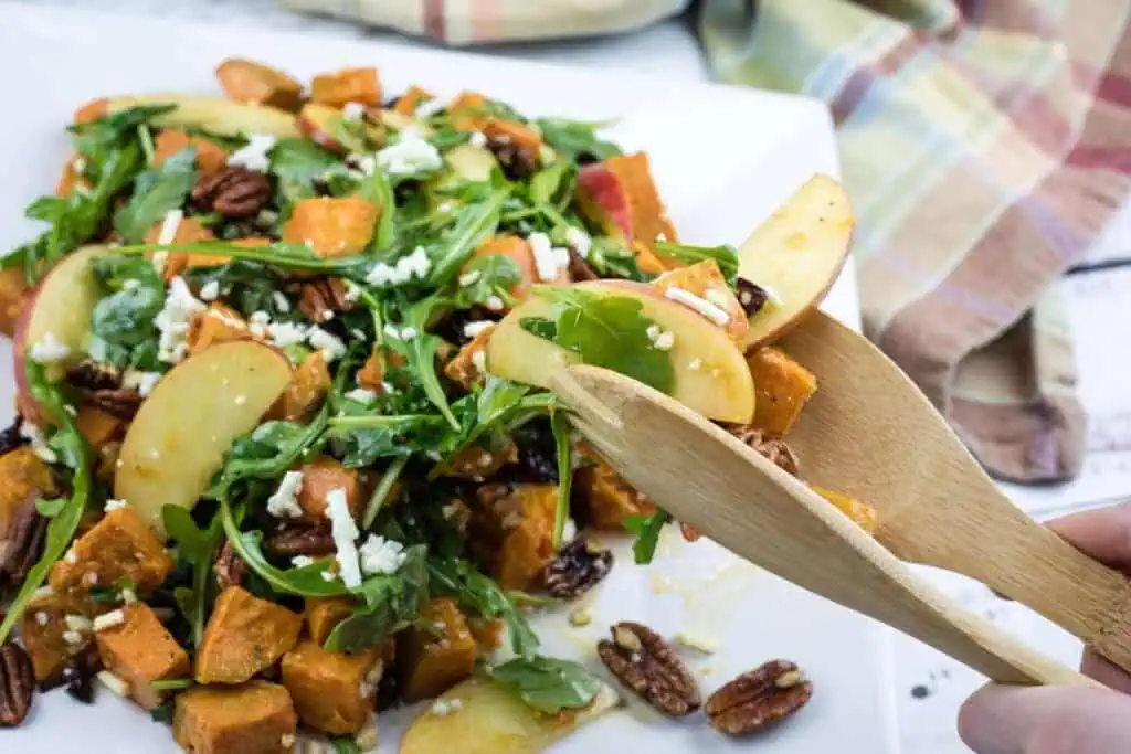 A plate of salad with arugula, sliced apples, sweet potatoes, pecans, and crumbled cheese, being served with wooden utensils.