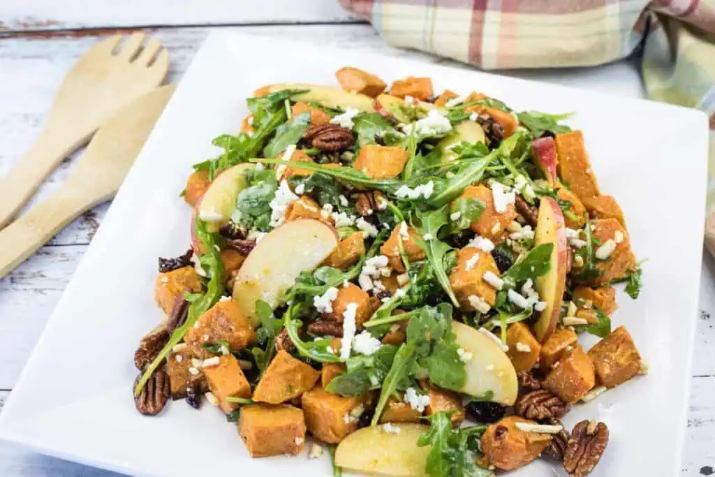 A salad with roasted sweet potatoes, apple slices, arugula, pecans, raisins, and crumbled cheese on a square white plate. Wooden utensils and a plaid napkin are nearby.