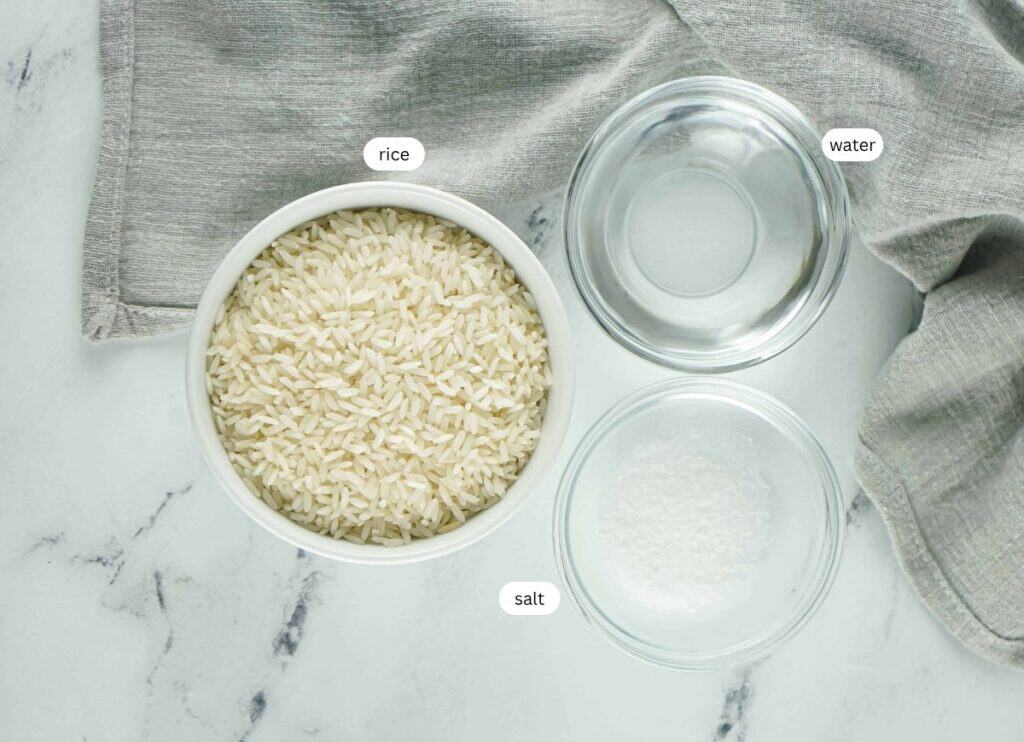 A bowl of uncooked rice, a bowl of water, and a bowl of salt are arranged on a marble surface next to a gray cloth.