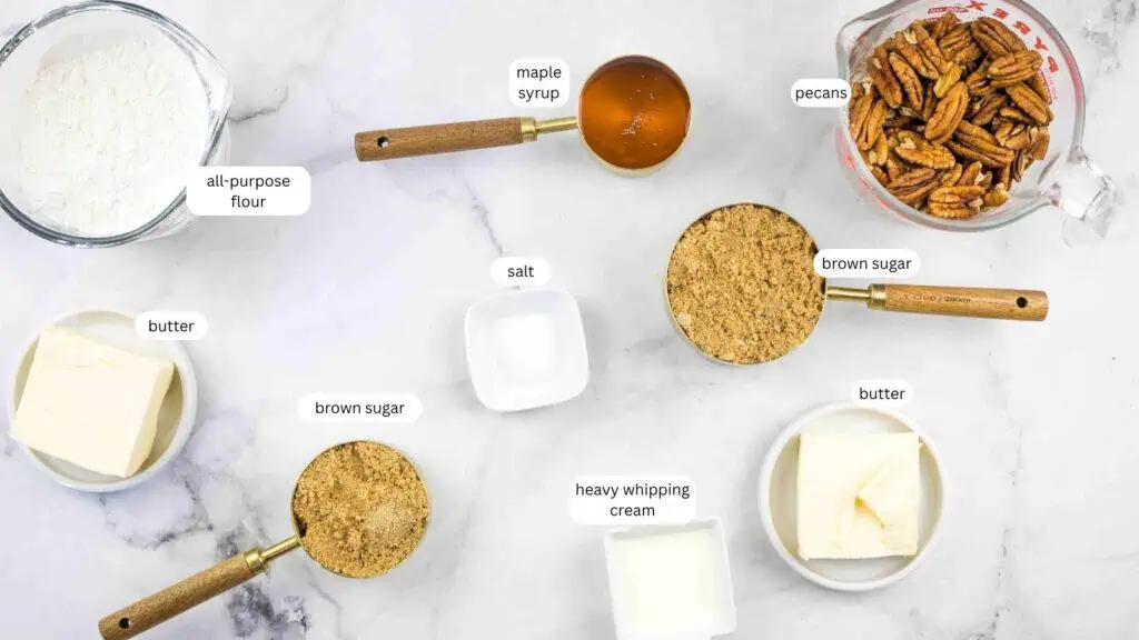 Top-down view of baking ingredients on a marble surface, including flour, butter, brown sugar, salt, maple syrup, heavy whipping cream, and pecans. Each item is labeled.