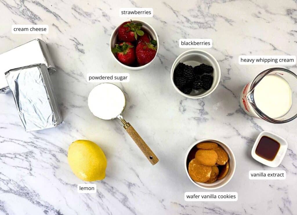 Various cheesecake ingredients are arranged on a marble surface, including cream cheese, powdered sugar, lemon, strawberries, blackberries, heavy whipping cream, wafer cookies, and vanilla extract.