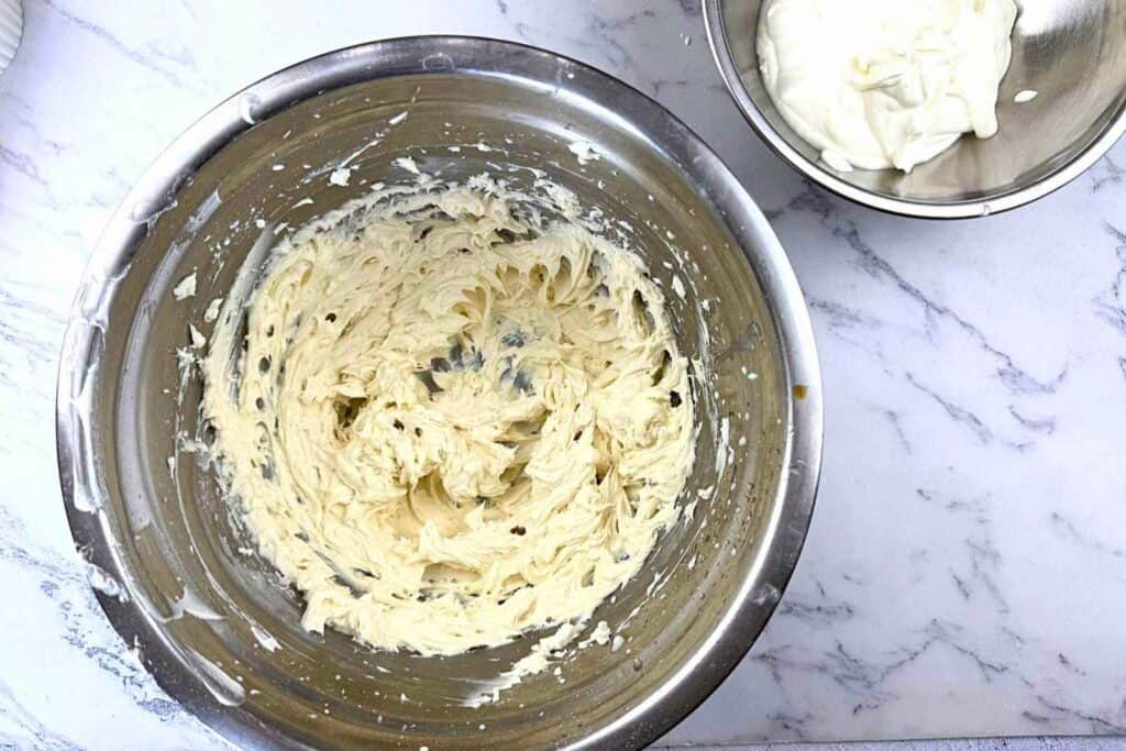 A large metal bowl with creamy, whipped batter sits on a marble surface next to a smaller bowl containing more cream.