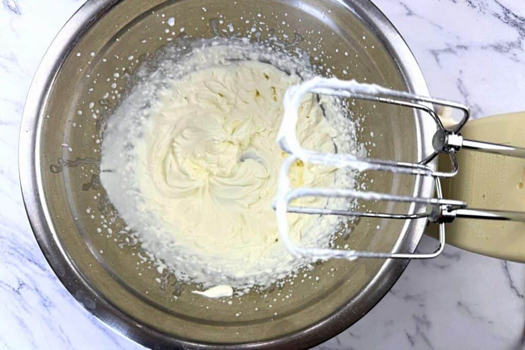 A metal mixing bowl with whipped cream and an electric hand mixer with beaters coated in whipped cream on a marble countertop.