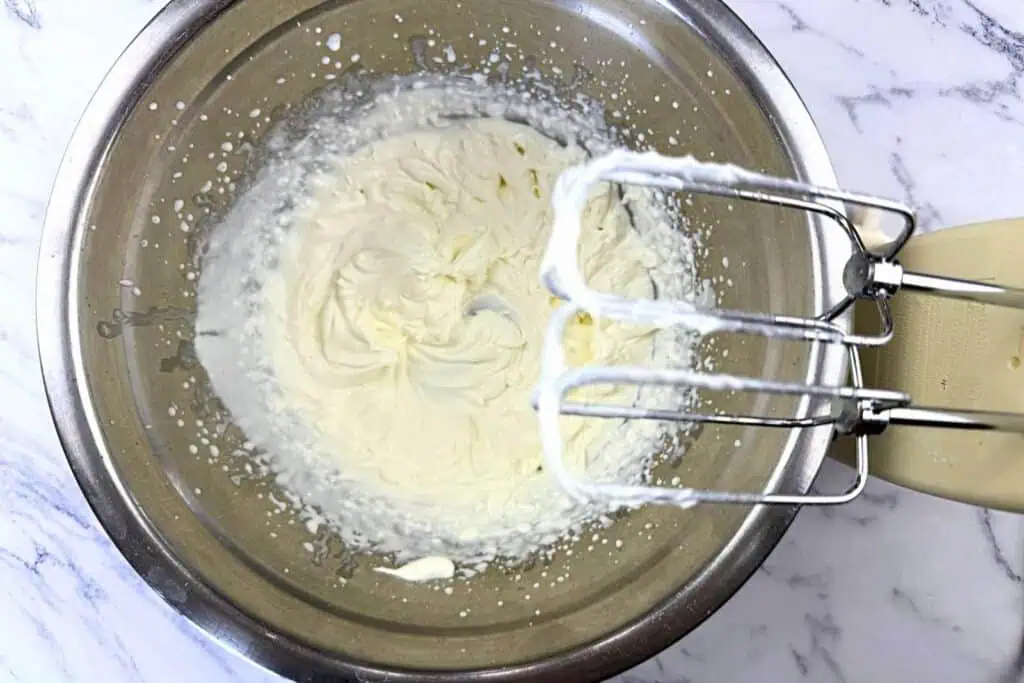 A metal mixing bowl with whipped cream and an electric hand mixer with beaters coated in whipped cream on a marble countertop.