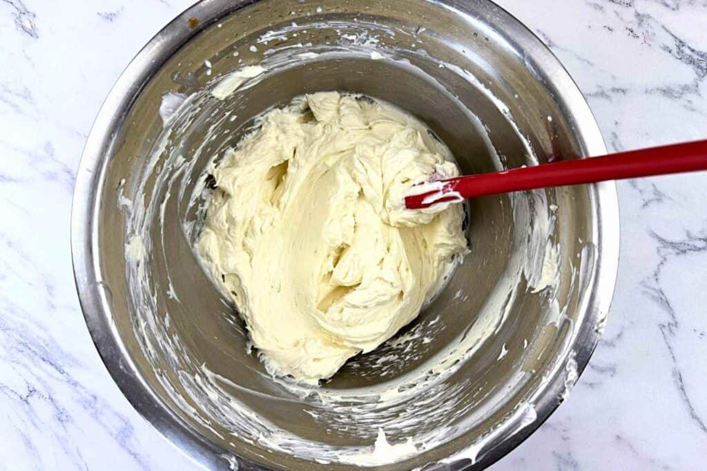 A metal mixing bowl with thick, creamy white batter being stirred by a red spatula, on a marble countertop.