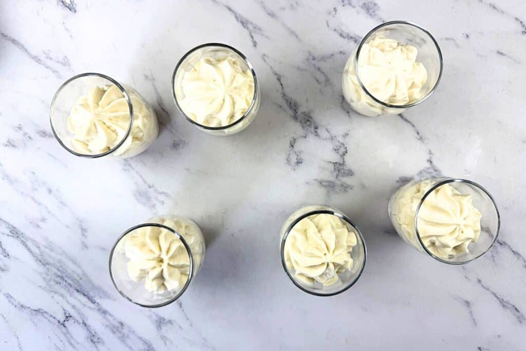 Six glasses filled with piped white cream are arranged on a white marble surface, viewed from above.