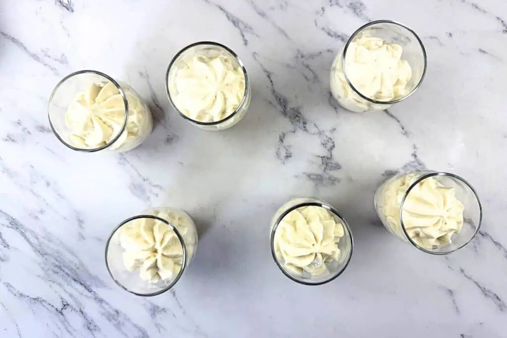 Six glasses filled with piped white cream are arranged on a white marble surface, viewed from above.