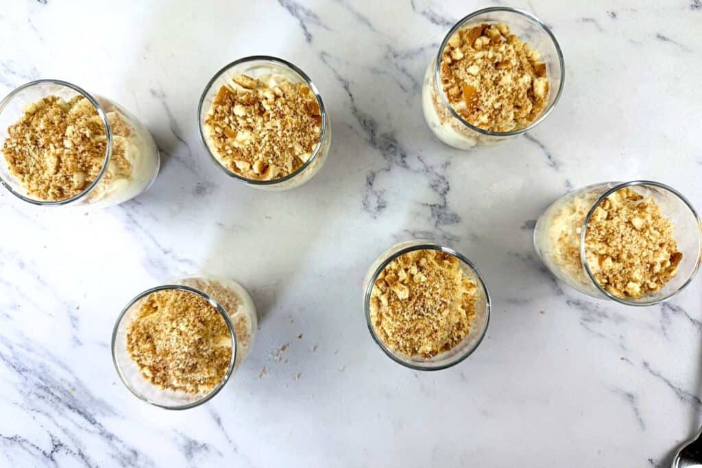 Six glasses filled with a dessert topped with crushed nuts or crumbs are arranged on a white marble surface.